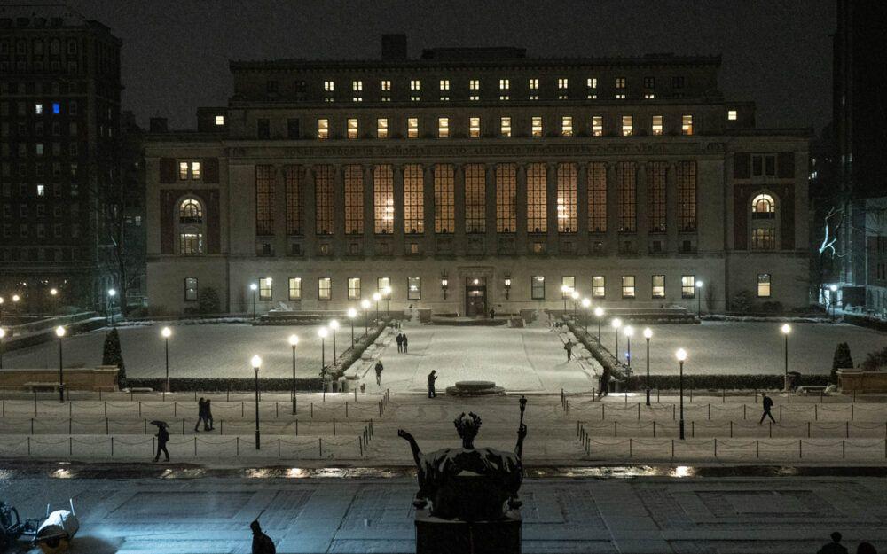 Illuminated building at night with snow-covered grounds and people walking along lit pathways.