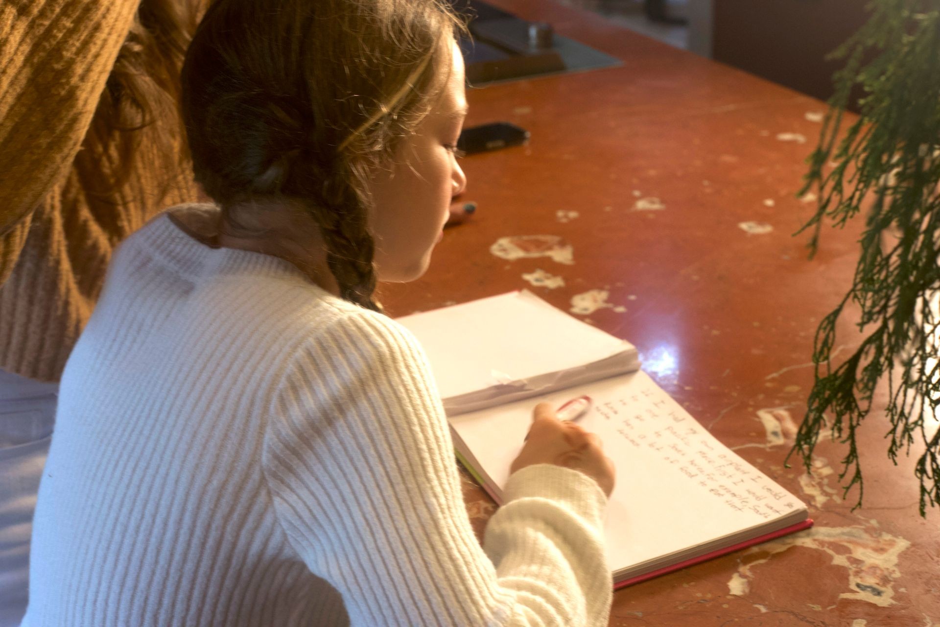 Child in a white sweater writing in a notebook at a brown stone table with greenery.